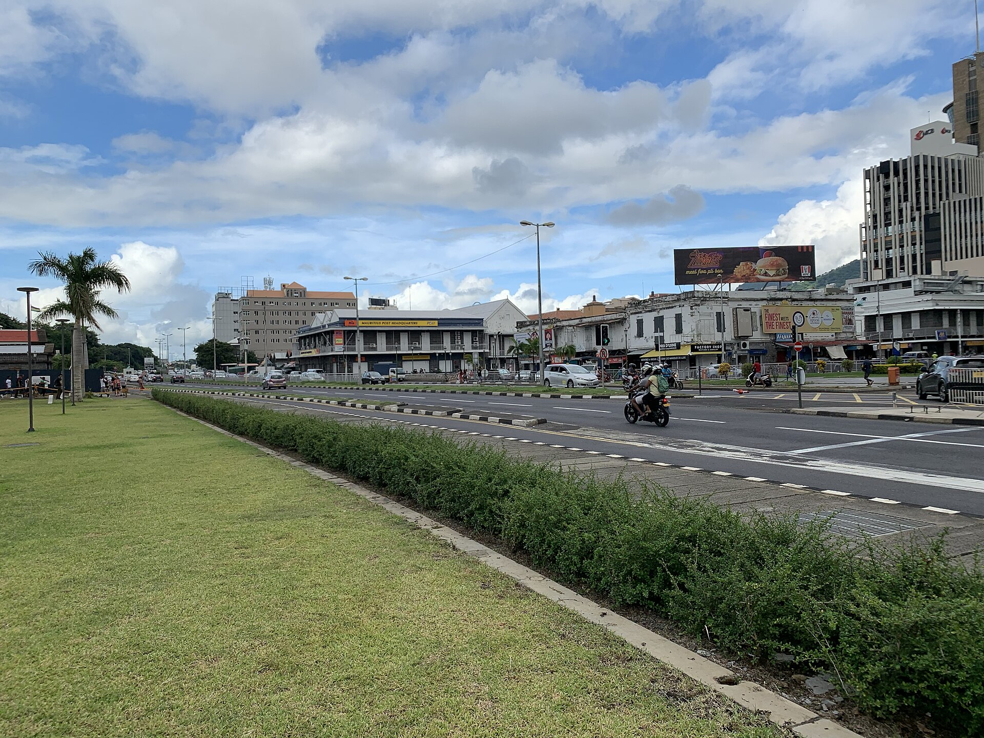 A wide main road in central Port Louis with a motorbike carrying two riders and cars beyond a grass verge